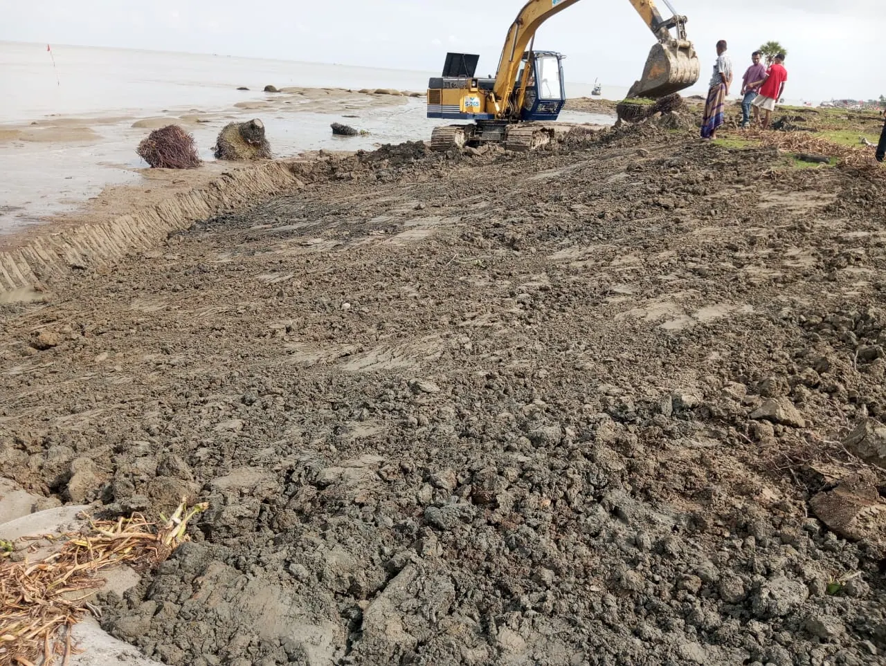 Yellow excavator grading earth on a riverbank slope beside the Meghna River under an overcast sky