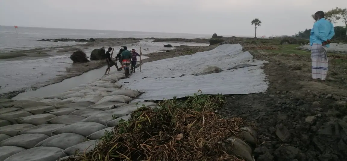 Workers laying geotextile sheet and geobags at the water's edge on an eroded riverbank at dusk