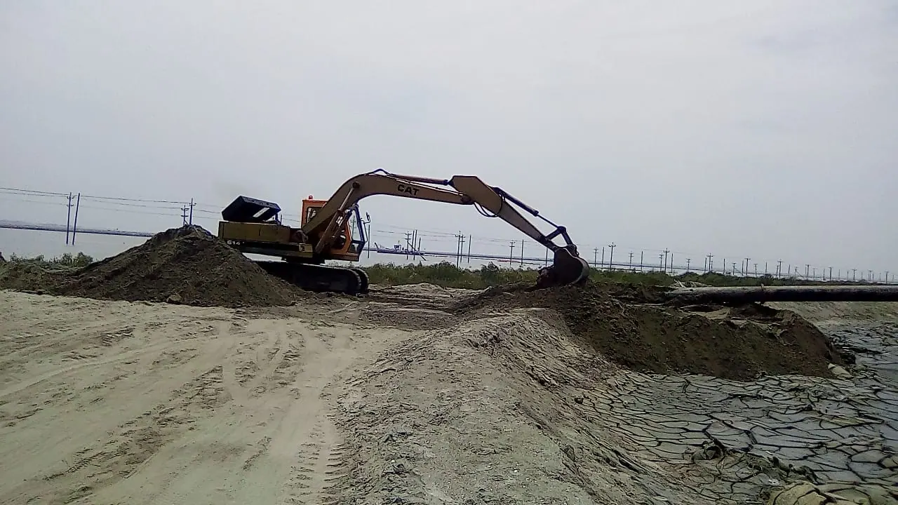 CAT excavator on a sand embankment beside a dredge discharge pipeline, river and power lines behind