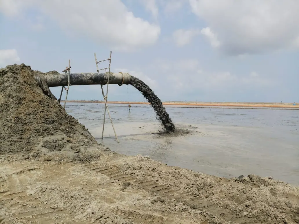 Dredge discharge pipe on bamboo supports pouring slurry into a reclamation pond under a cloudy sky