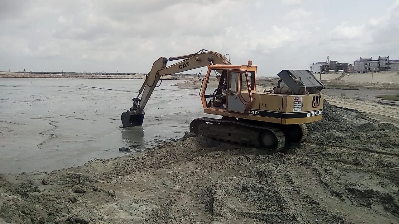 CAT excavator digging at the water's edge on freshly placed dredged sand, buildings under construction behind