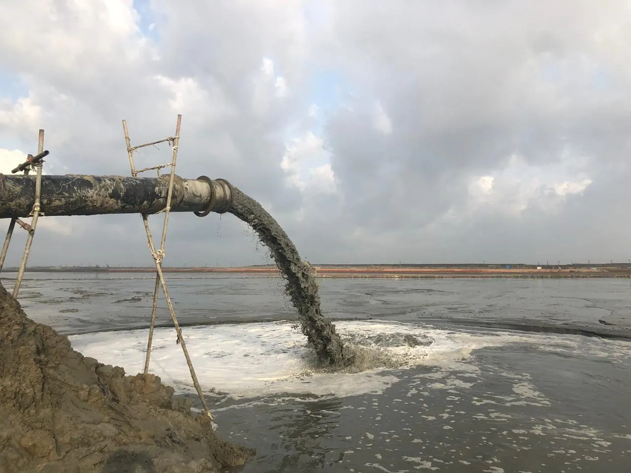 Dredge discharge pipe on bamboo supports pouring a thick stream of slurry into a foaming reclamation pond