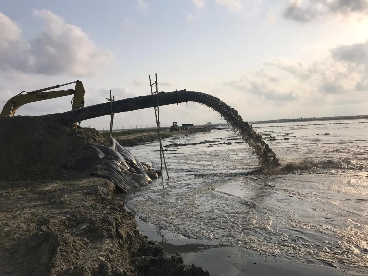 CAT excavator boom beside a dredge pipeline arcing slurry into the reclamation pond, backlit cloudy sky