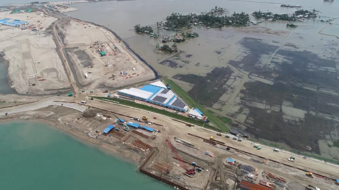 Wide aerial of reclaimed coastal land at Matarbari with a blue-roofed accommodation camp, haul roads and sheet pile works