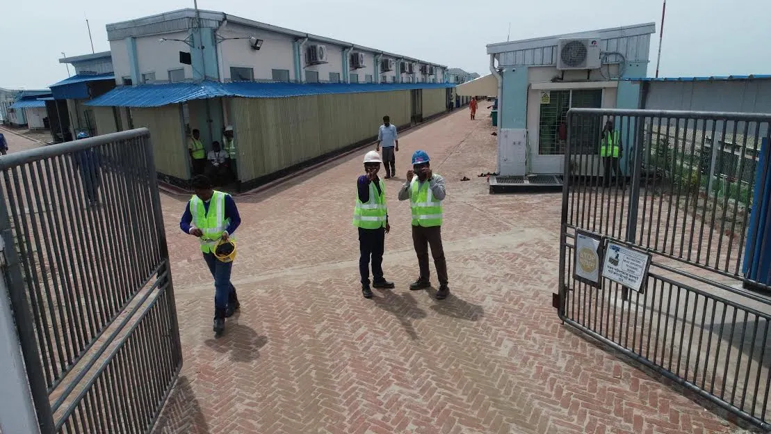 Workers in hi-vis vests and hard hats walking through the open steel gate of a brick-paved accommodation camp