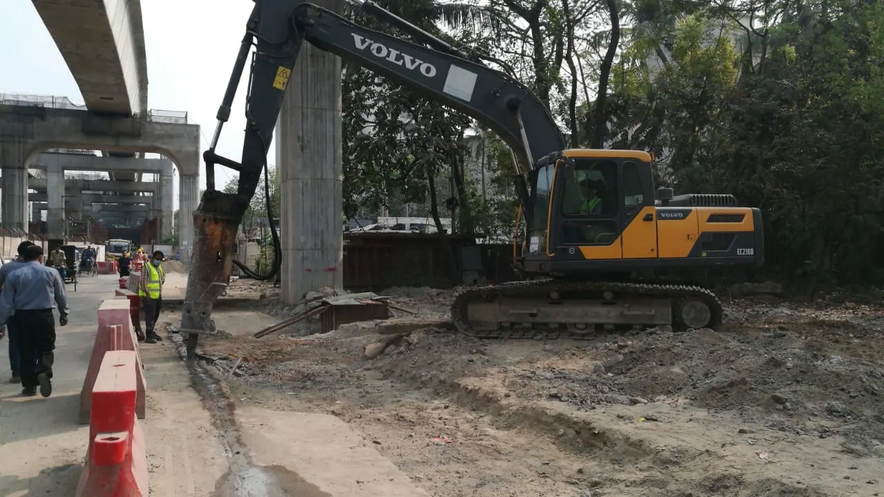 Volvo excavator with hydraulic breaker breaks old pavement next to a Metro Rail pier, elevated viaduct behind