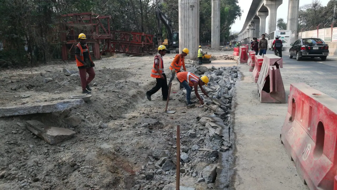Workers in orange vests break up old concrete pavement by hand beside red barriers under the Metro Rail viaduct