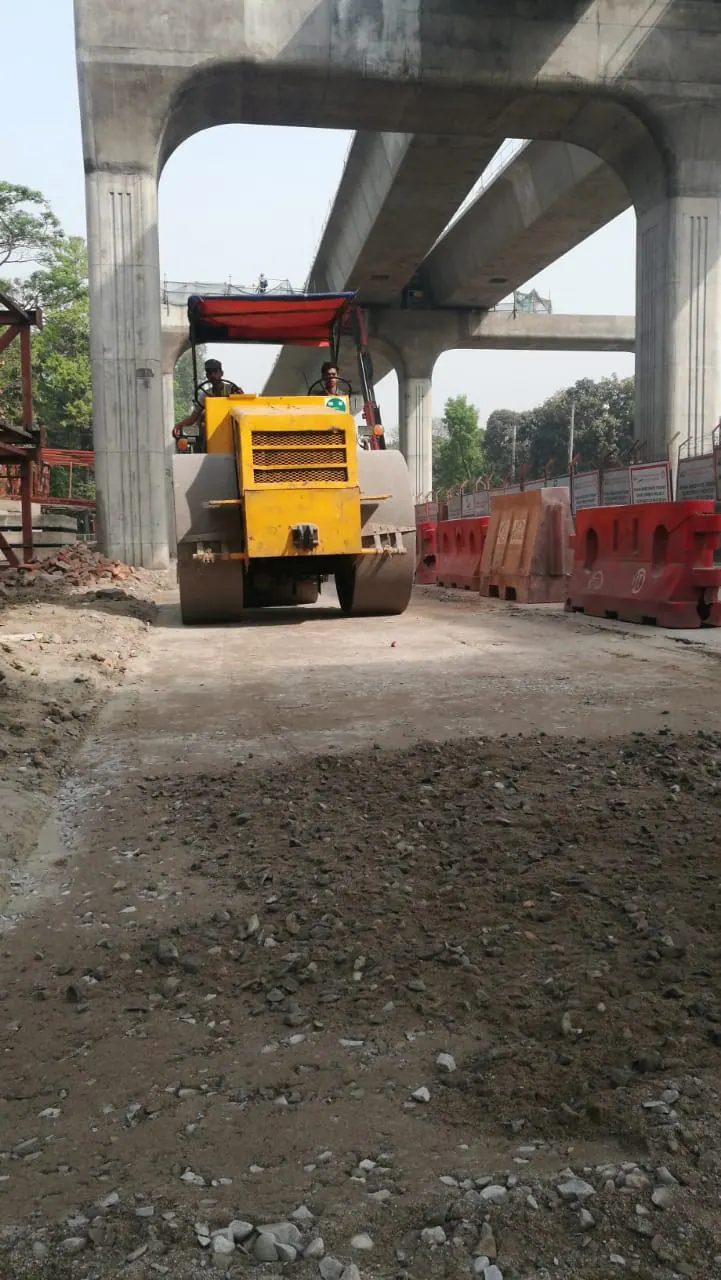 Yellow tandem road roller with canopy compacts base course under a Metro Rail viaduct arch, red barriers at right