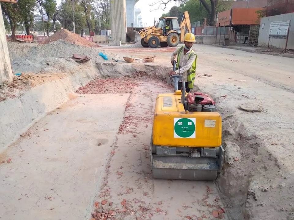 Operator guides a walk-behind vibratory roller over brick aggregate sub-base in a trench, backhoe loader behind