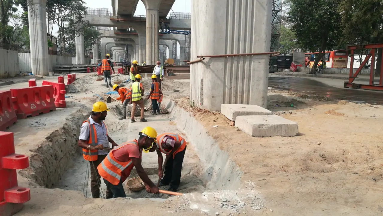 Workers with shovels and baskets excavate a pavement trench beside a Metro Rail pier, more crew and viaduct behind