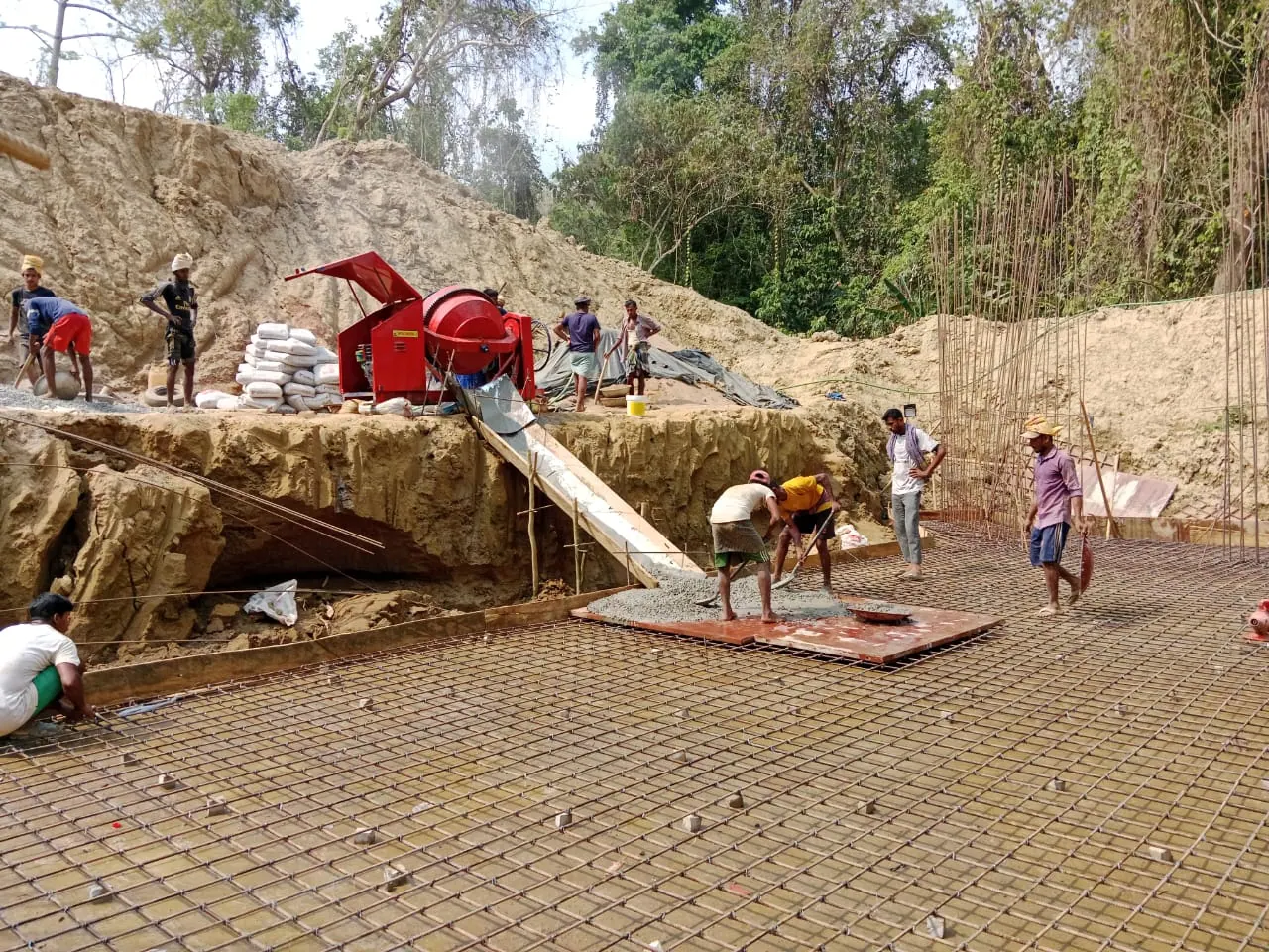 Workers spreading concrete from a mixer chute onto rebar mesh of a culvert base slab in a hillside excavation