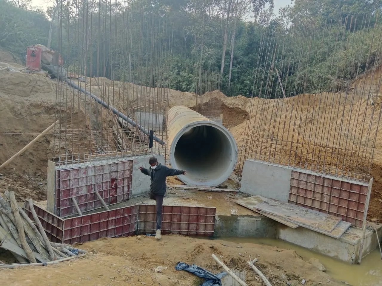 End view of an RCC pipe culvert barrel flanked by rebar cages and red steel shuttering, worker on formwork