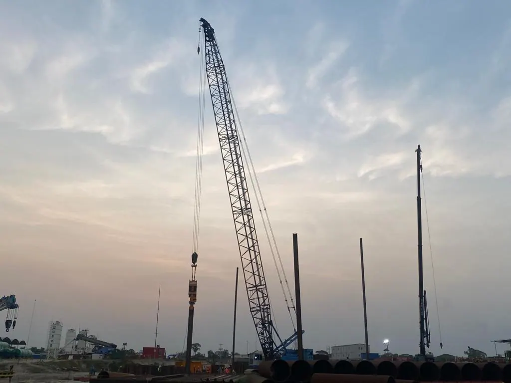 Crawler crane with vibro hammer driving a steel pile at dusk, stacked pipe piles in the foreground