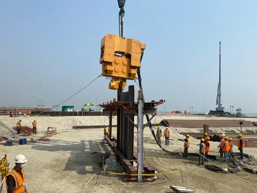 Vibro hammer suspended from a crane above a steel sheet pile guide frame, workers handling hydraulic hoses