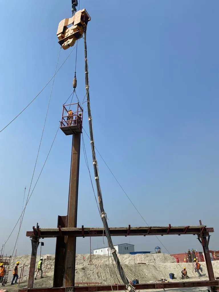 Workers in a red man basket atop a tall steel pile below a crane-held vibro hammer, blue sky behind