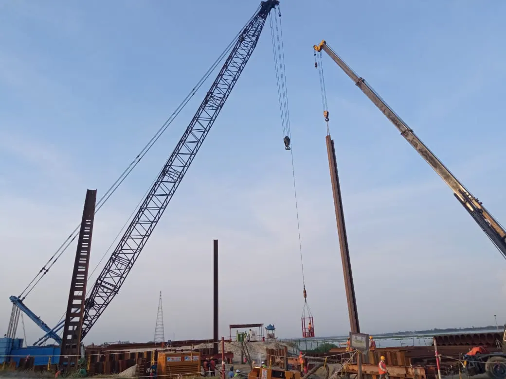 Crawler crane and telescopic mobile crane lifting a steel pile upright against a blue sky at a riverside site