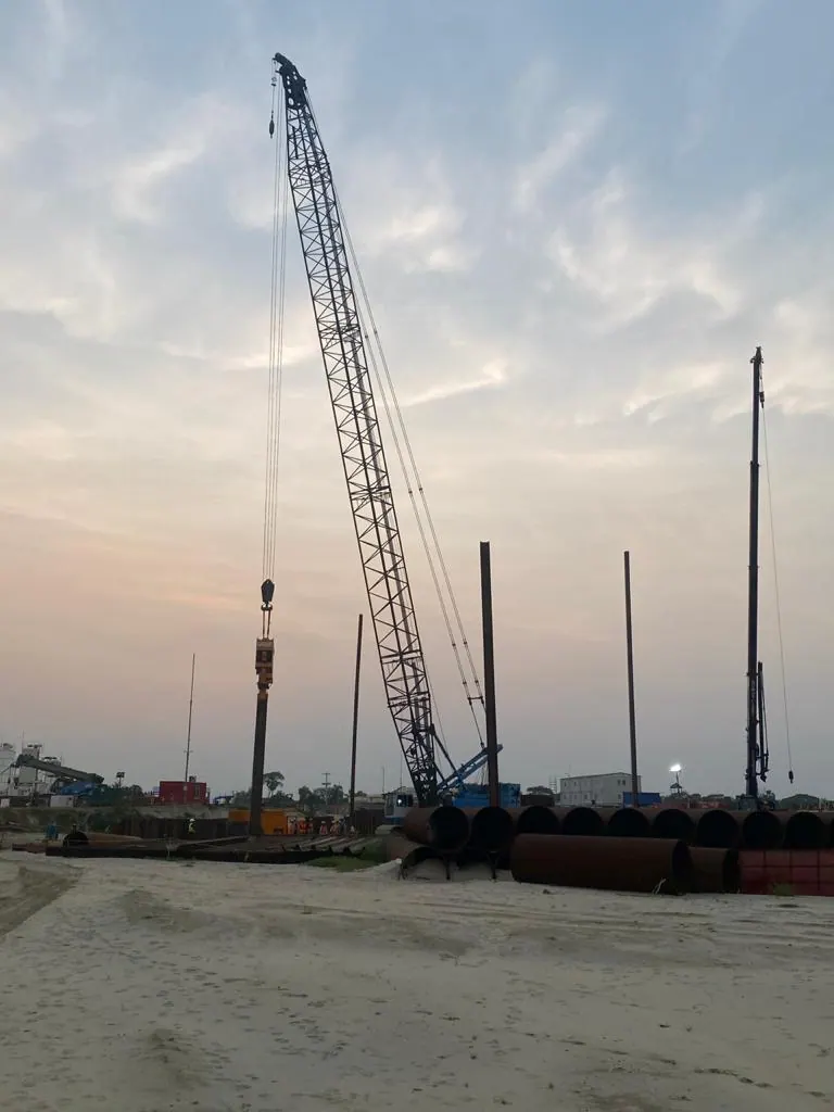 Crawler crane with vibro hammer driving a pile at dusk, stacked steel pipes and sandy ground in the foreground
