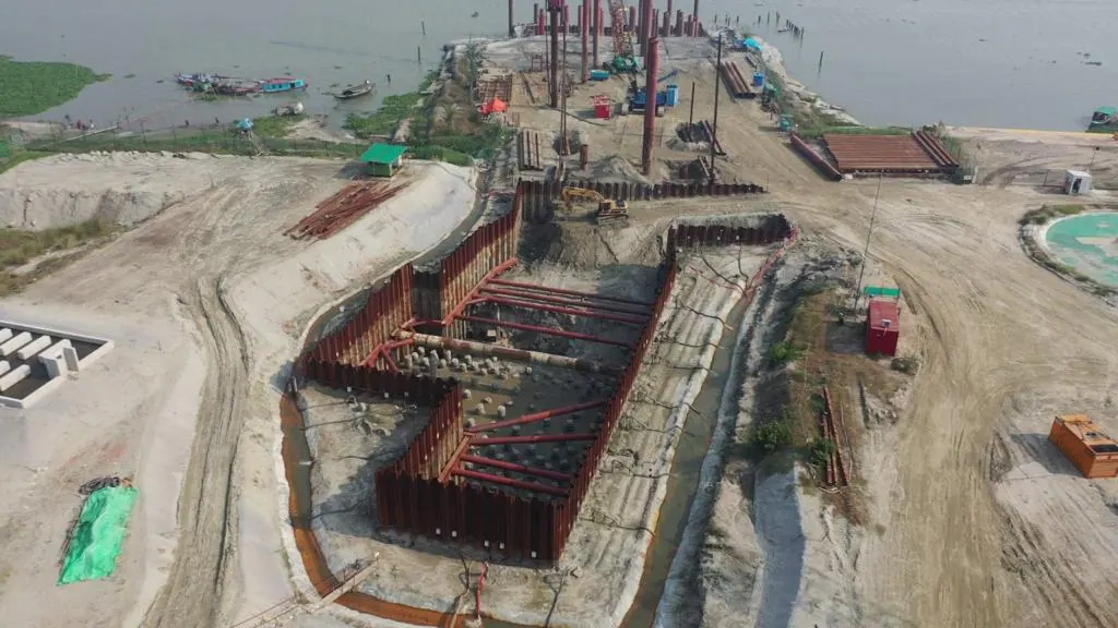 Aerial view of a sheet pile cofferdam excavation with pipe piles and red pipework beside a river with boats