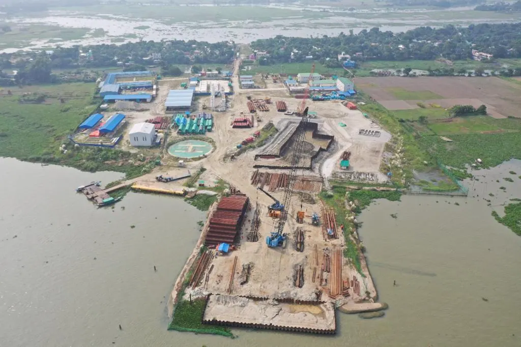 Aerial view of a riverside construction site with site offices, stacked steel pipes, cranes and a cofferdam