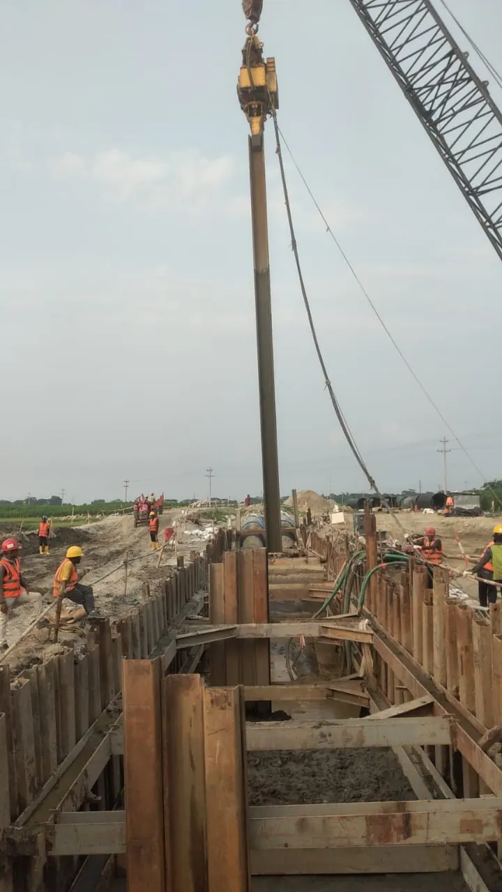 Crawler crane driving a sheet pile along a braced trench for a pipeline, workers in orange vests on both sides